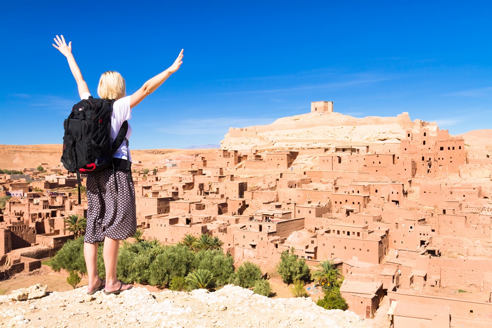 Adventurous-woman-arms-reised-in-front-of-Ait-Benhaddou-fortified-city-kasbah-or-ksar-along-the-former-caravan-route-between-Sahara-and-Ma.jpg